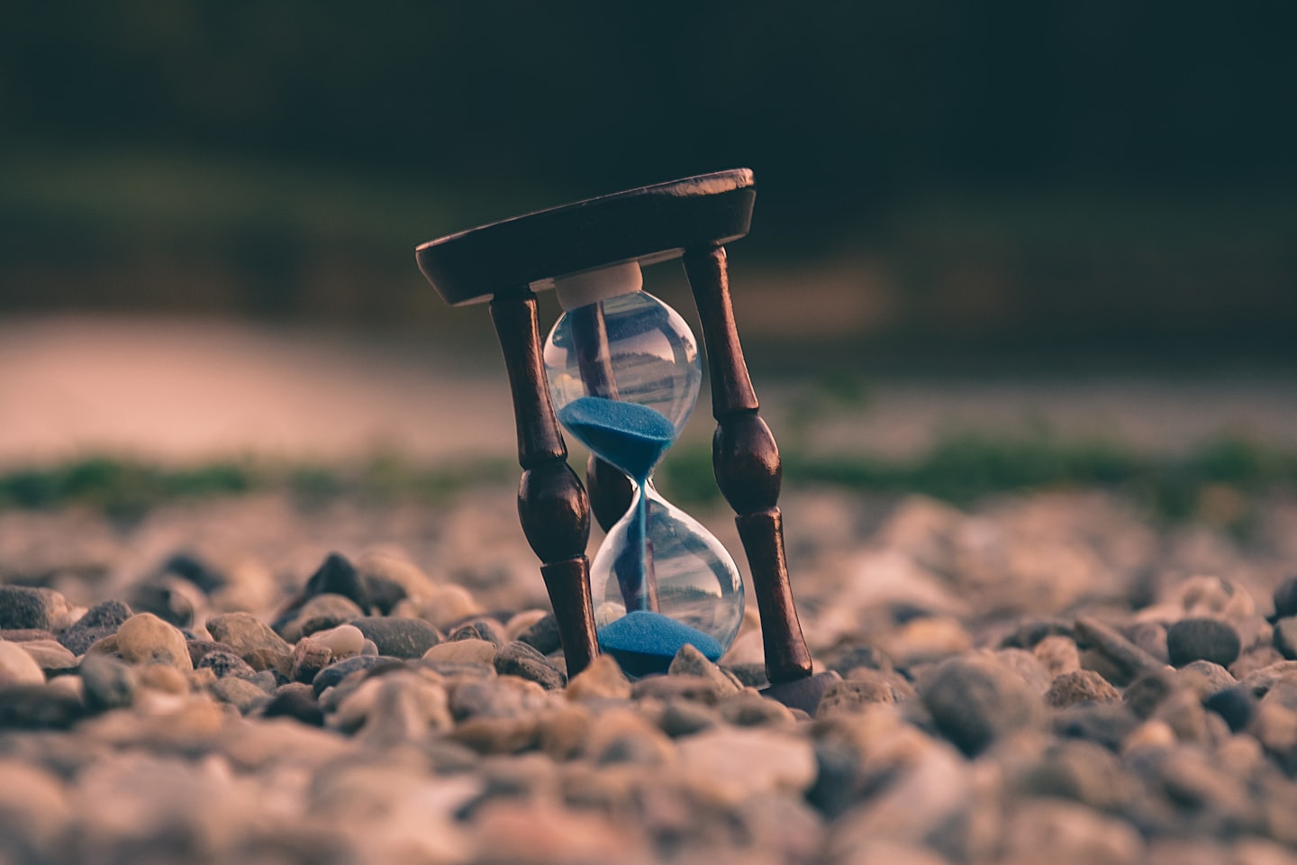 AGWAT hourglass with blue sand resting on pebbles at sunset, symbolizing time, focus, and taking action today.