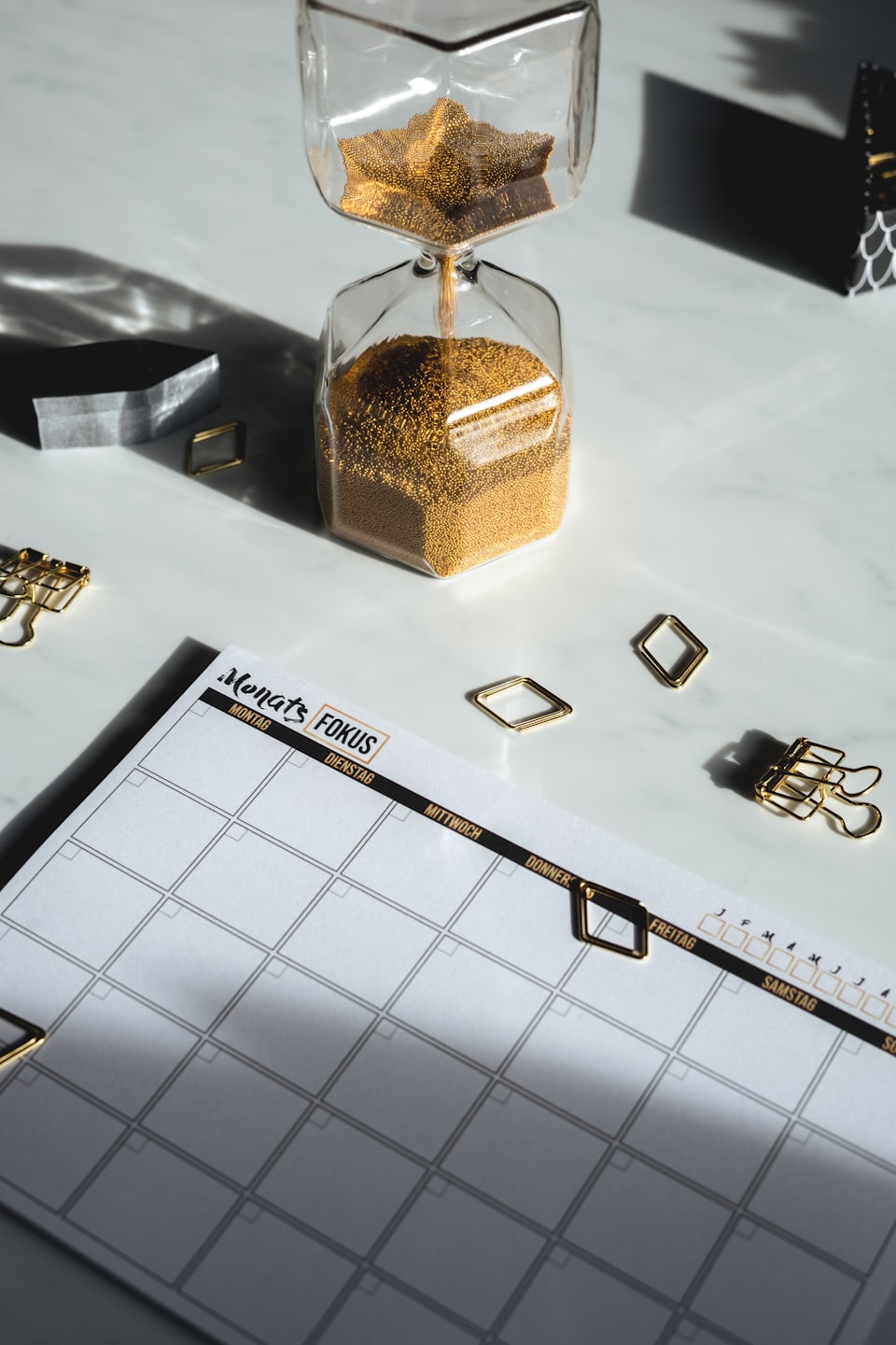 AGWAT golden hourglass beside a monthly focus calendar on a marble desk, symbolizing time management and deliberate action.